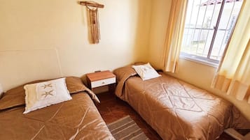 A room with two beds, a small wooden nightstand, a wall-mounted macrame decoration, and a window with curtains.