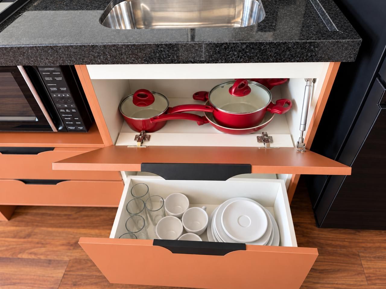 A kitchen with a built-in microwave, a sink, and a drawer with red pots and white dishes.