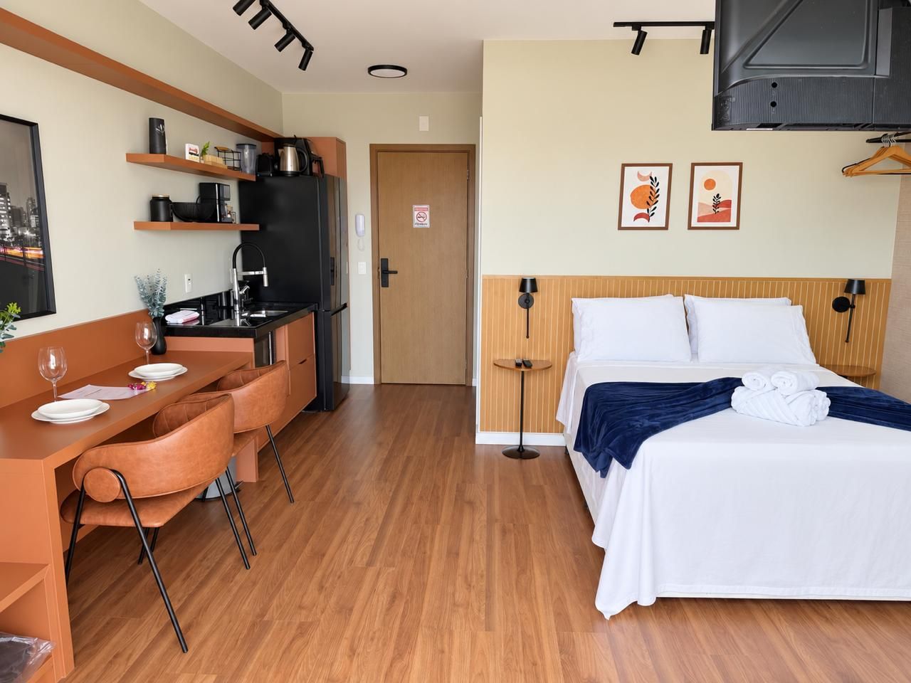 A compact hotel room with a kitchenette, a bed with white linens, a small desk with a chair, and a television mounted on the wall.