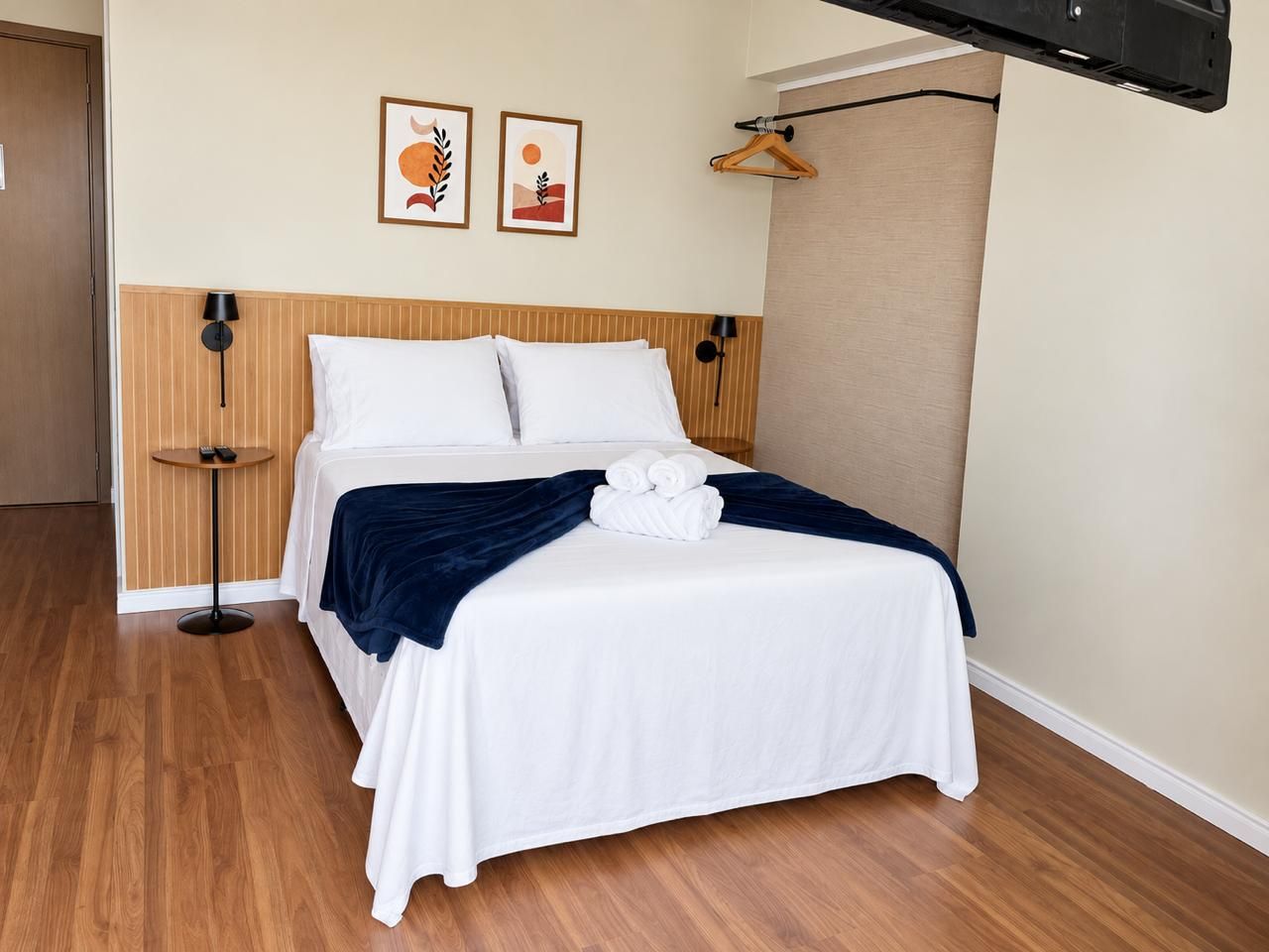 A hotel room with a wooden floor, a bed with white linens, a dark blue throw blanket, and two framed pictures on the wall.