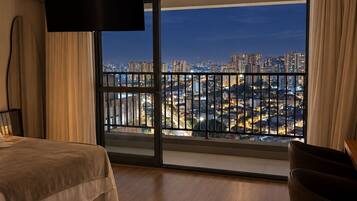 Habitación de hotel con una cama grande, televisión, escritorio y vista a un paisaje urbano desde un balcón.