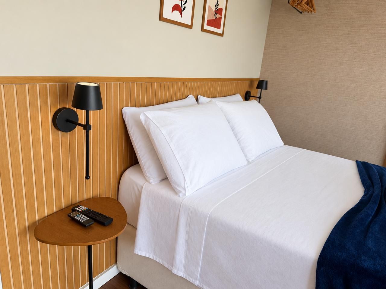 Quarto de hotel com cama, mesa de cabeceira com controle remoto e luminária na parede.