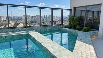 Piscina no terraço com vista para a cidade, rodeada por guarda-vidro e fachada de um edifício moderno.
