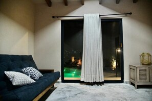 A dark blue sofa with patterned pillows, a sliding glass door with a white curtain, and a small wooden side table with a woven basket. - Cuernavaca Downtown Golf Club Boutique House with a heated pool (Cuernavaca)