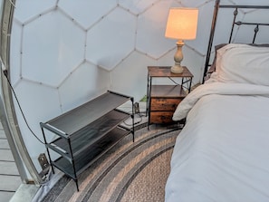 A bed with a white comforter, a bedside table with a lamp, a small wooden chest, and a metal shelf unit. - Taurus | Black Lake Glamping Dome (Hammond)