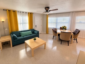 Un salon comprenant un canapé, une table basse, une table à manger avec des chaises, un ventilateur de plafond et une porte.