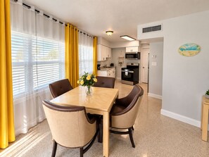 Un comedor con una mesa y sillas, una cocina al fondo y una ventana con cortinas.