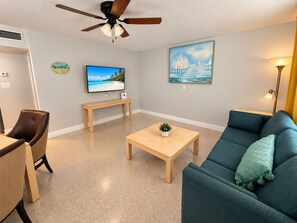 A living room with a ceiling fan, a television, a painting, a sofa, a coffee table, and a bench. - Cozy 1BR Beach Cottage, Steps from Gulf, Dogs Welcome (Redington Shores)