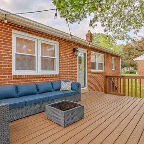 Een overdekt terras met een blauwe bank en rieten poef, een bakstenen huis met ramen met witte kozijnen, en een houten omheining.