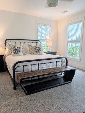 Cama com estrutura de metal, roupa de cama branca e almofadas estampadas. Tem uma mesa de cabeceira com luminária e vista para o verde pela janela.