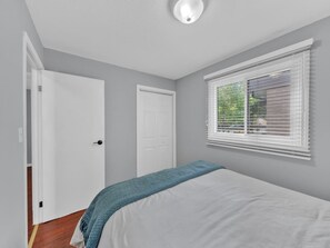 A bedroom with a bed, a window with blinds, a door, and a ceiling light. - 4BR Home | Fenced Yard & 2-Car Parking (Windsor)