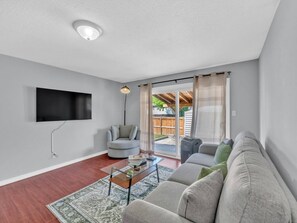 A living room with a flat-screen TV mounted on the wall, a grey sofa, a round grey armchair, and a glass coffee table with a book and remote. - 4BR Home | Fenced Yard & 2-Car Parking (Windsor)