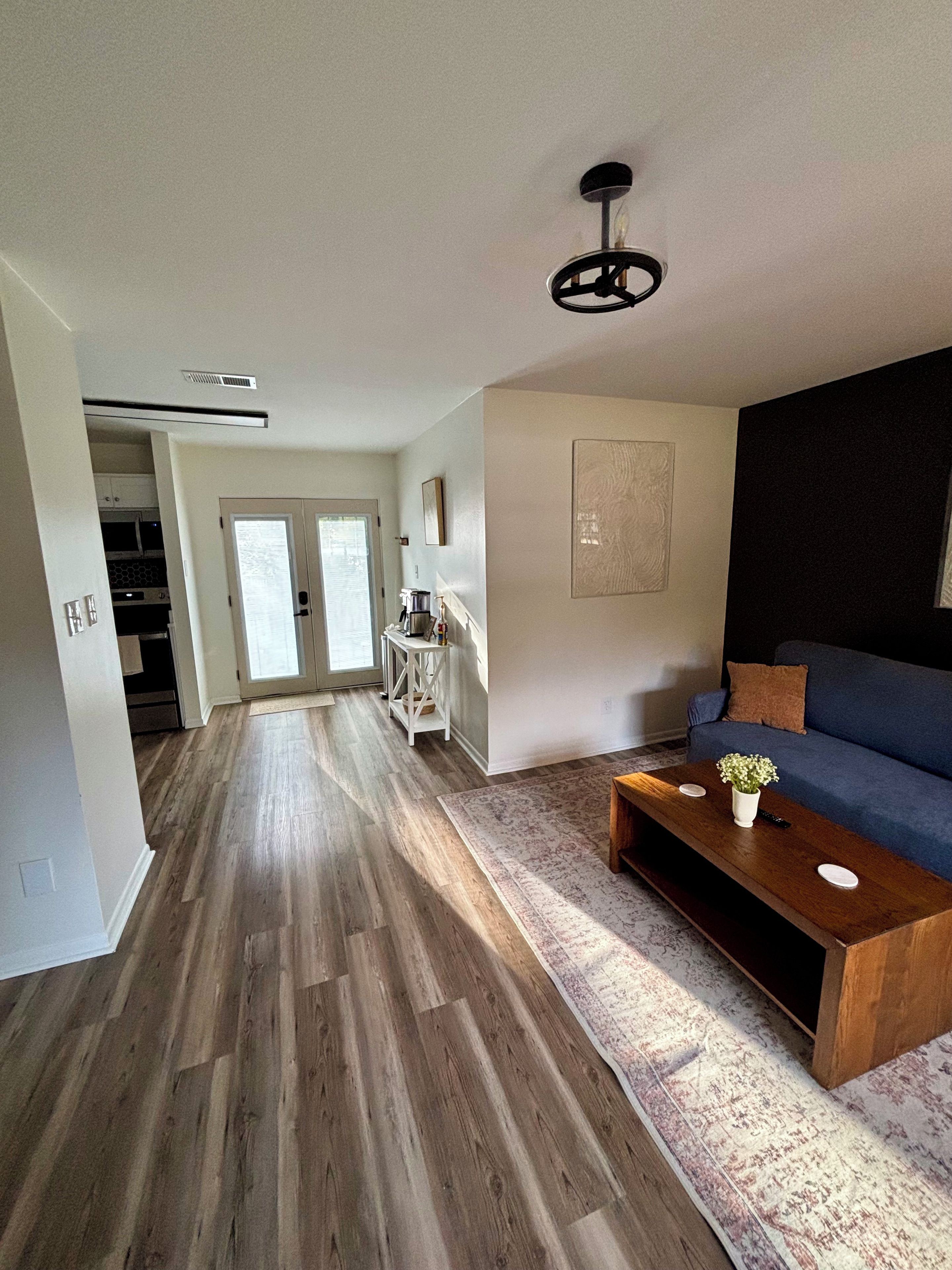 A modern living room with a wooden floor, a blue sofa, a coffee table with a vase, and a kitchen in the background.