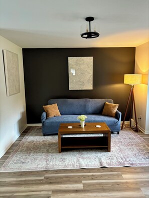 Ein Wohnzimmer mit einem blauen Sofa, einem Holztisch, einer Stehlampe und einem gemusterten Teppich.