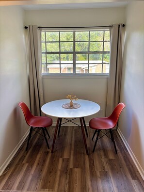 Een kamer met een ronde tafel, twee stoelen, een raam met gordijnen en uitzicht op groen.