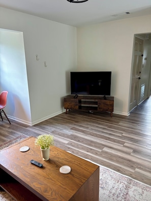 Ein modernes Wohnzimmer mit einem Flachbildfernseher auf einem Holzbodenständer, einem Couchtisch mit Fernbedienungen und einer Blumenvase, sowie einem Flur, der zu einer Tür führt.