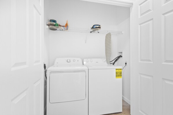 Uma lavanderia com máquina de lavar e secadora de carregamento frontal, prateleira com produtos de limpeza e mangueira de ventilação.
