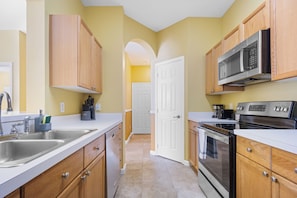 A kitchen with wooden cabinets, stainless steel appliances, and a white door in the background. - Condo 2BR en Windsor Palms cerca de Disney (Kissimmee)