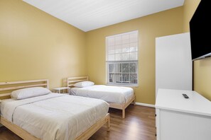 Quarto com duas camas de solteiro, escrivaninha branca, televisão e janela com persianas.