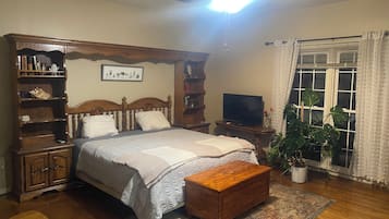 A bedroom with a bed, wooden headboard, bedside tables, a television, a ceiling fan, a window with curtains, and a potted plant.
