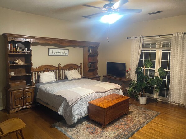Un dormitorio con cama, cabecera de madera, mesitas de noche, televisor, ventilador de techo, ventana con cortinas y una planta en maceta.