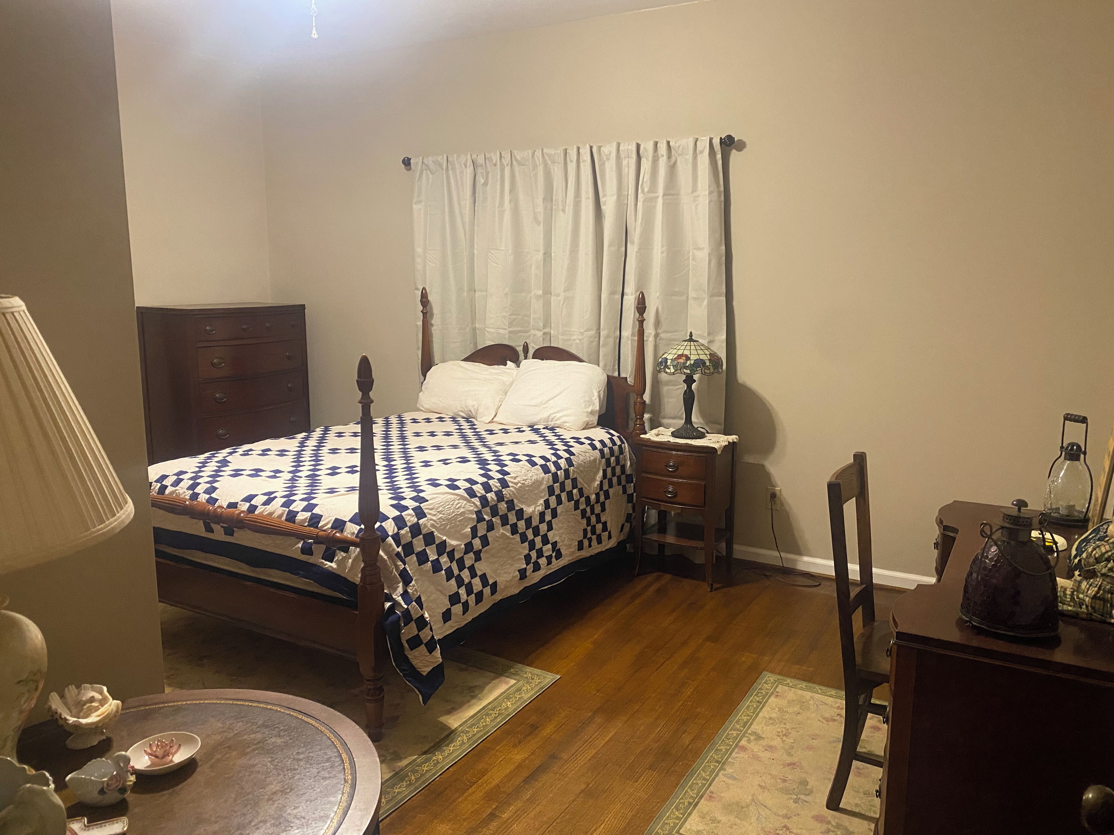 Een slaapkamer met een hemelbed, een nachtkastje met lamp, een houten dressoir en een stoel.