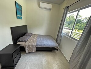 A bedroom with a bed, a nightstand, an air conditioning unit, and a window with a view of greenery. - Pet Friendly • 2BR/1BA apartment close to the beach (Jacó)