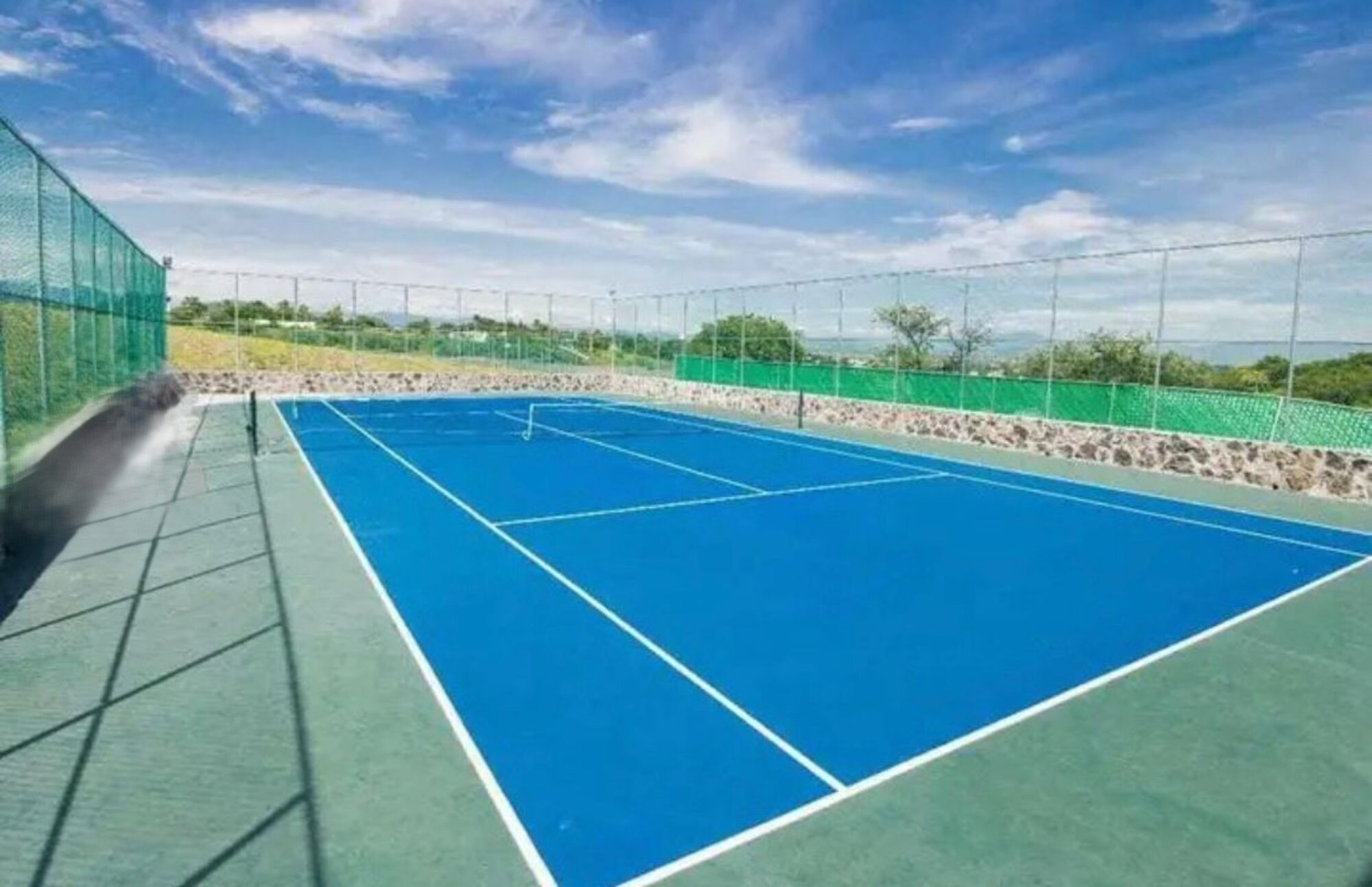 Una cancha de tenis al aire libre con superficie verde y azul, rodeada por una cerca verde y un muro de piedra.