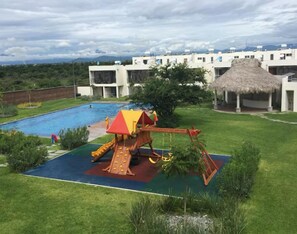 Piscina com escorregador e estrutura de telhado de palha, cercada por gramado e edifícios modernos.
