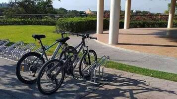 Un pavillon couvert avec un toit de chaume, un espace de stationnement pour vélos équipé de supports, et un chemin piétonnier dallé.