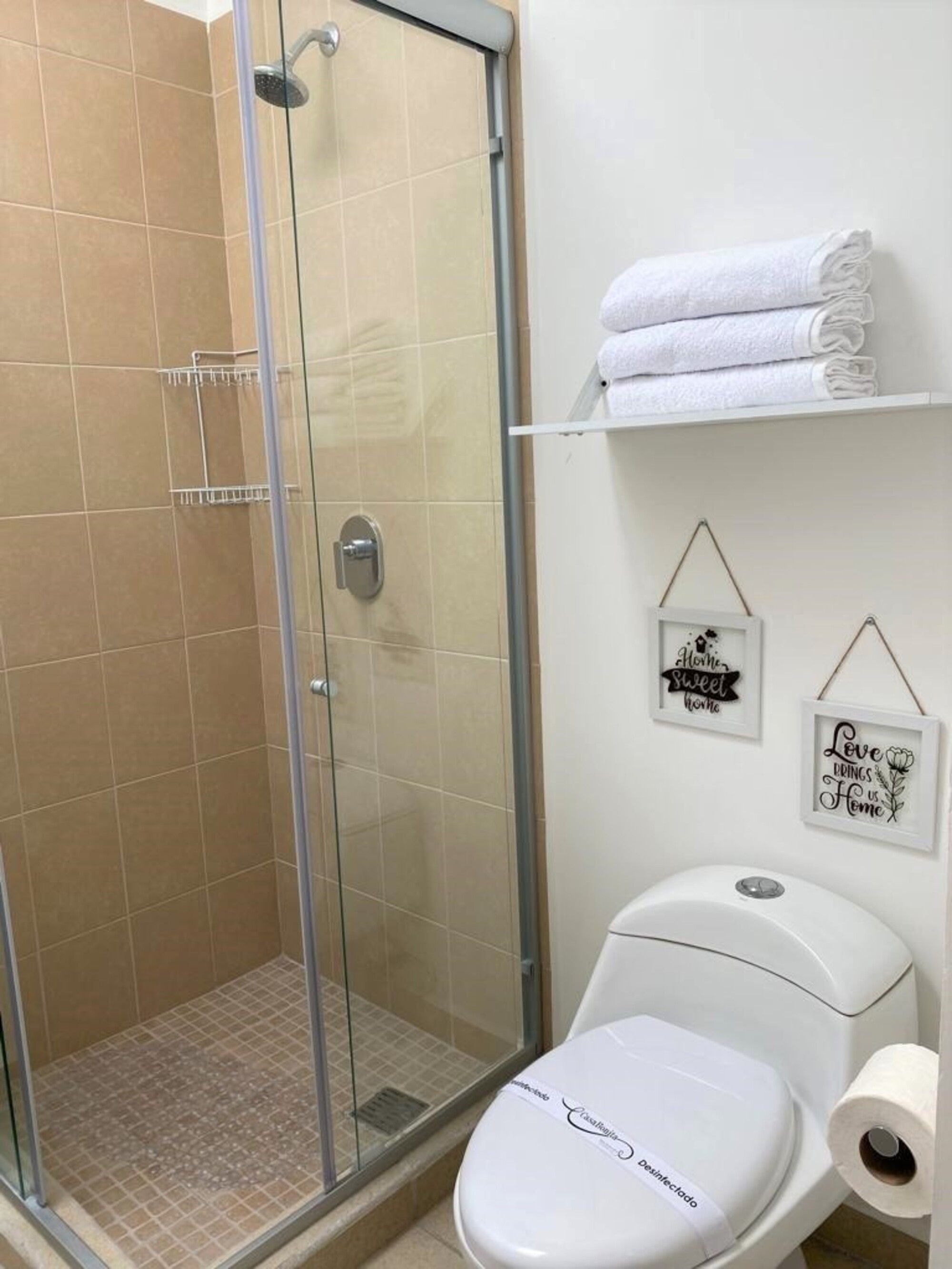 Un baño con ducha de vidrio, un inodoro con un rollo de papel higiénico y dos letreros enmarcados en la pared.