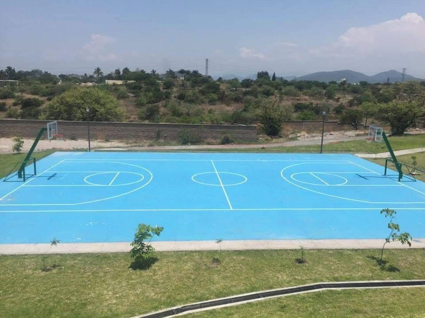 Una cancha de baloncesto al aire libre con dos aros, rodeada de césped y árboles, con un paisaje montañoso al fondo.