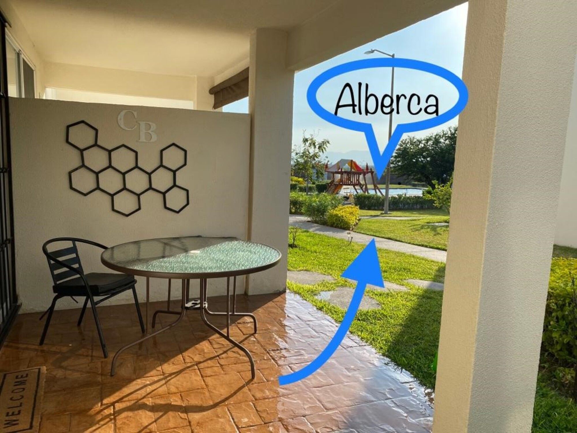 A patio with a glass table and chairs, a 'WELCOME' mat, hexagonal wall art, and a view of a playground and greenery.
