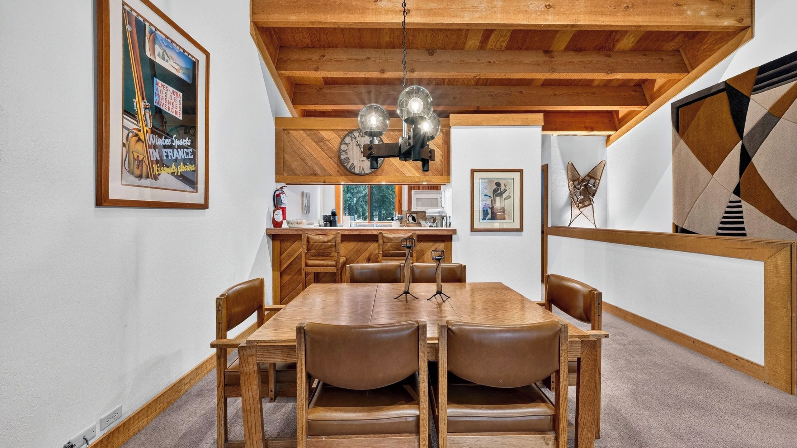 A dining area with a wooden table and chairs, a hanging light fixture, and a kitchen in the background.