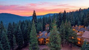 A lodge with multiple wooden cabins surrounded by dense forest, set against a backdrop of a mountain range and a colorful sky. - Northstar Pines by Avantstay Pine-view Deck Shared Pool + Spa Steps to Northstar Shuttle (Truckee)