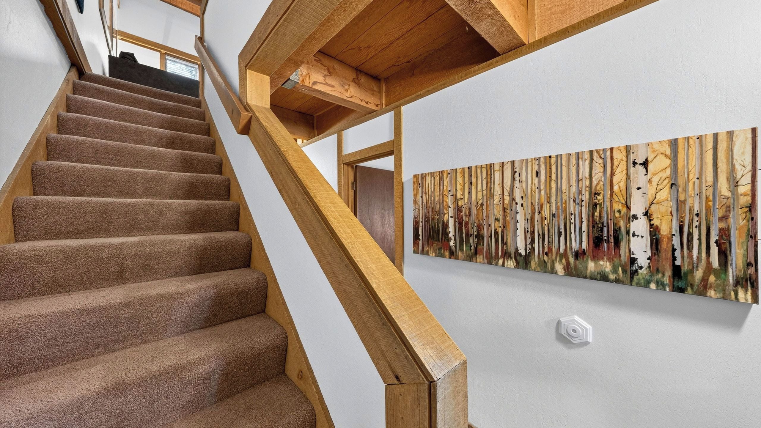 A staircase with carpeted steps and wooden handrails leading to a room with a white wall and a painting of trees.