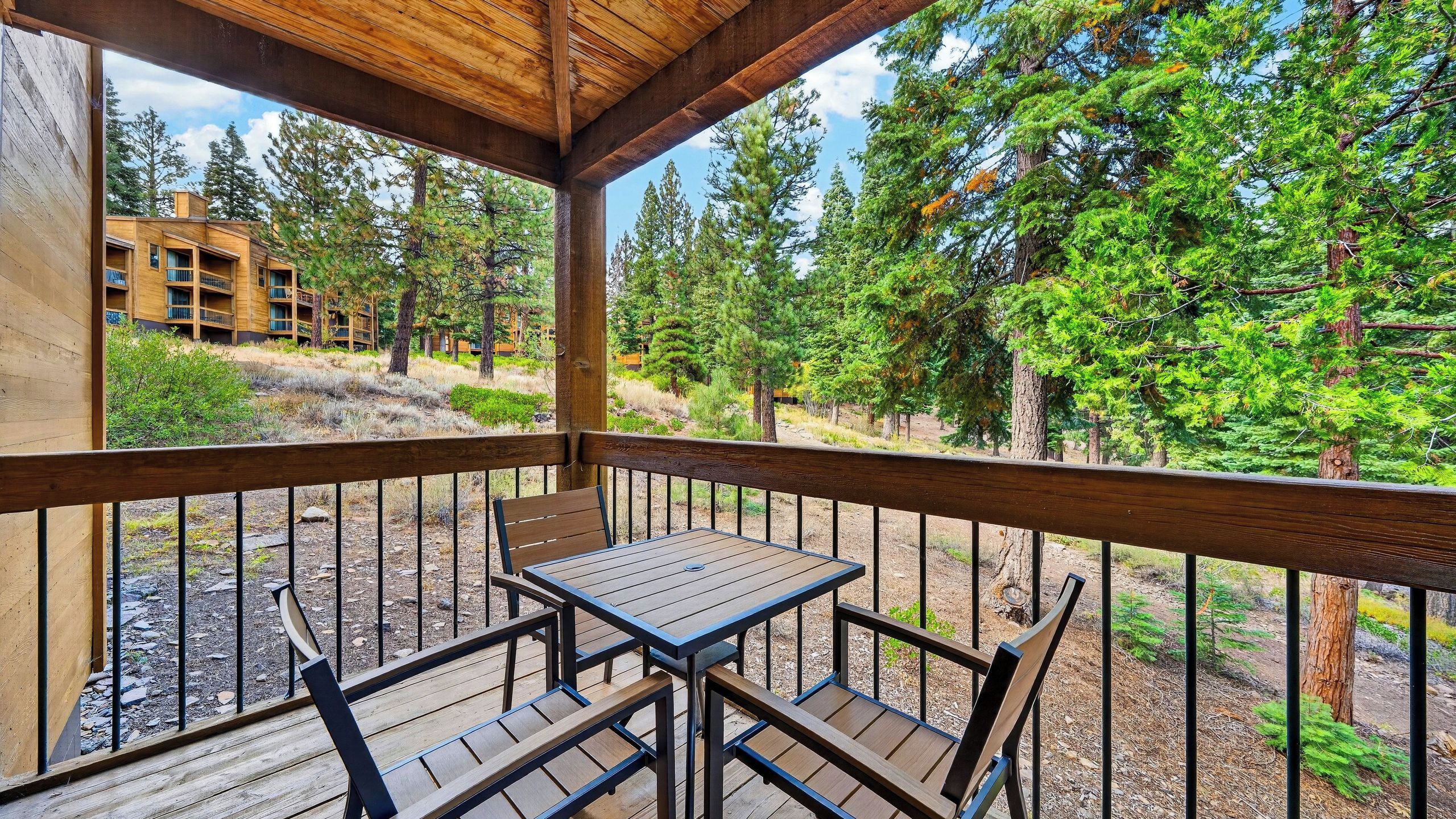A covered outdoor area with a table and chairs, surrounded by a railing, overlooking a forested area with a building in the background.