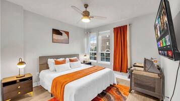 A hotel room with a bed, a desk, a chair, a ceiling fan, a window with curtains, and a wall-mounted TV.