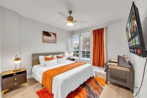 Een hotelkamer met een bed, een bureau, een stoel, een plafondventilator, een raam met gordijnen en een aan de muur bevestigde televisie.