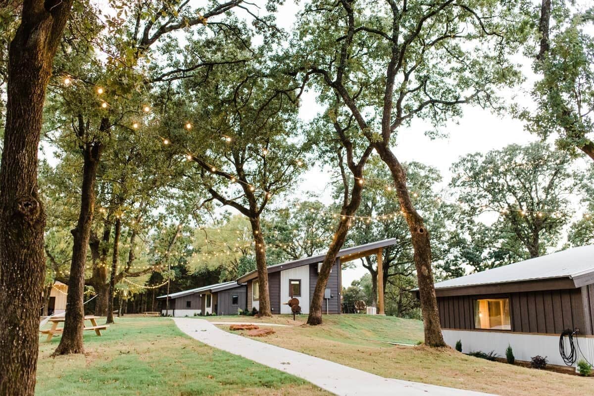 A row of cabins with a pathway leading through a wooded area, string lights overhead, and picnic tables.