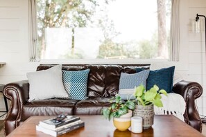 A brown leather sofa with patterned cushions, a wooden coffee table with books and a plant, and a window with a view of trees. - Texas Tiny Cabins #6 – Serene with Hilltop Views (Grayson County)