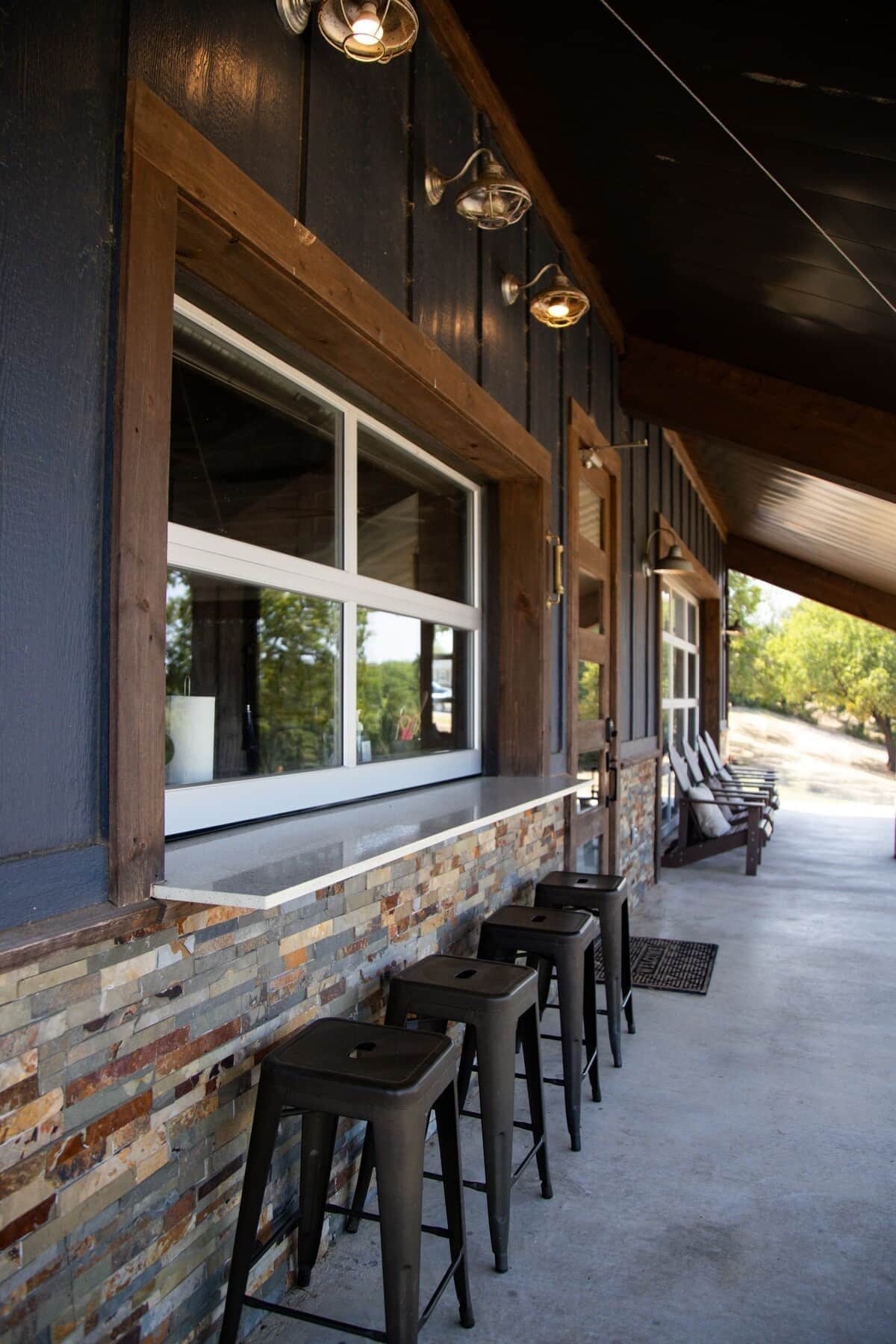 A covered outdoor seating area with a row of bar stools and a window overlooking a parking lot.