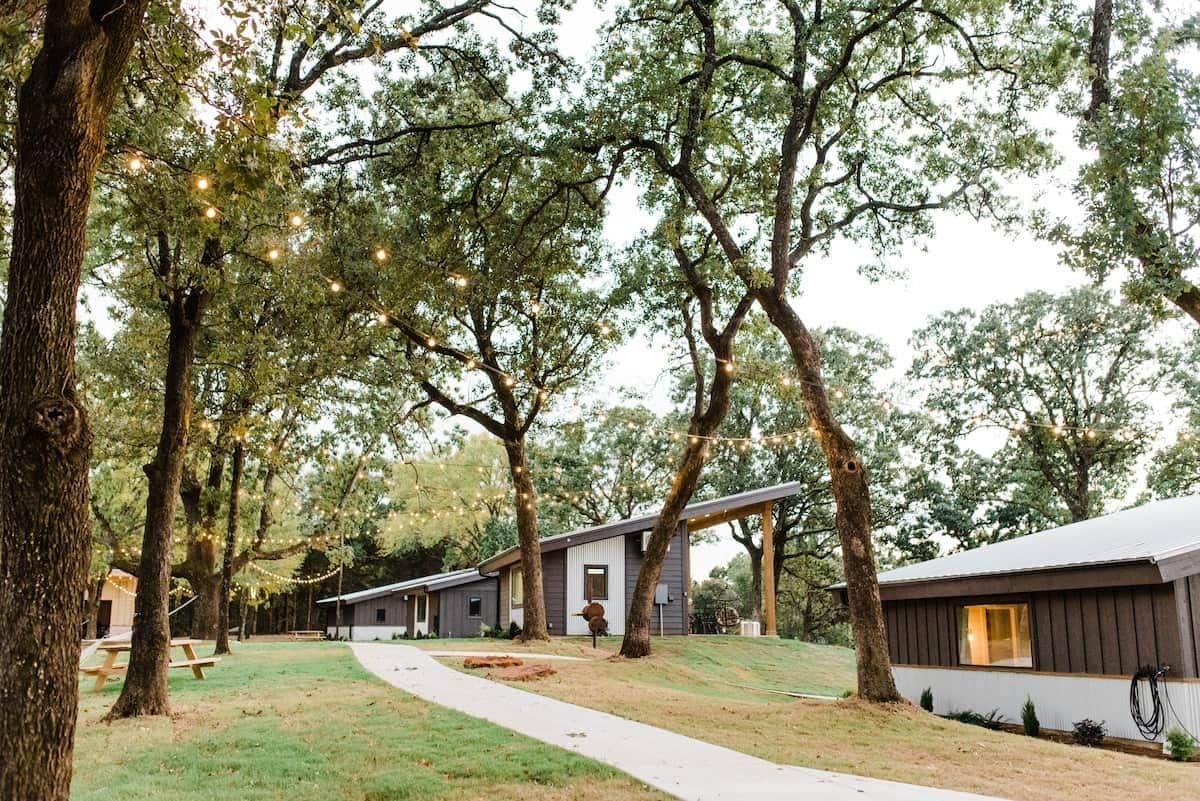 A row of cabins with a pathway leading through a wooded area, string lights overhead, and picnic tables.