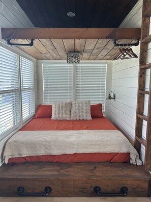 A bedroom with a wooden ceiling, a bed with red bedding, white pillows, and a window with blinds. - Tiny Home With The OK View (Grayson County)