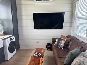 A living room with a flat-screen TV mounted on the wall, a brown leather sofa with decorative pillows, a wooden coffee table, and a washing machine in the corner. - Tiny Home With The OK View (Grayson County)