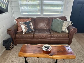 A brown leather sofa with patterned cushions, a wooden coffee table with remote controls, and a window with blinds. - Tiny Home With The OK View (Grayson County)