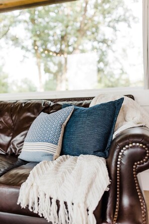A brown leather sofa with a white throw blanket and two pillows, one with a blue pattern and the other plain, positioned in front of a window with a view of trees. - Texas Tiny Cabins #2 – Peaceful Cabin, Scenic View (Grayson County)