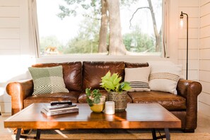 A brown leather sofa with patterned cushions, a wooden coffee table with books and a plant, and a view of trees through a window. - Texas Tiny Cabins #4 – Quiet Cabin on 40 Acres (Grayson County)