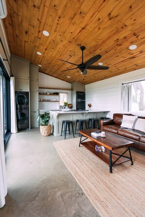 A modern kitchen with a central island, wooden ceiling, and a seating area with a leather couch and coffee table. - Texas Tiny Cabins #4 – Quiet Cabin on 40 Acres (Grayson County)
