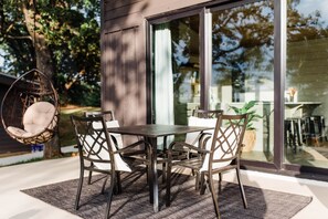 Uma área de refeições ao ar livre com mesa e cadeiras, uma cadeira suspensa e uma porta de vidro que dá para um jardim.